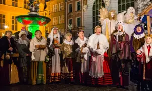 Grupa kobiet w strojach historycznych śpiewa kolędy. W tle iluminowana fontanna i zabytkowe kamienice.
