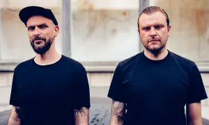 Dwóch mężczyzn w czarnych koszulkach. Jeden w czapce z daszkiem, drugi z zaczesanymi do tyłu włosami. Ich tatuaże sugerują, że tworzywo sztuczne za mało czasu na ich życie.
