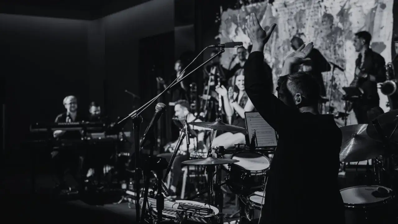 Muzycy na scenie, w tym perkusista z podniesionymi rękami, tworzą dynamiczną atmosferę. To kwintesencja polskiego jazzu.