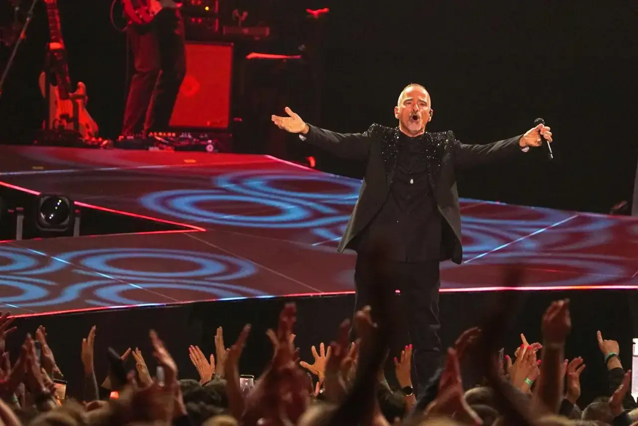 Eros Ramazzotti na scenie, w czarnym garniturze, z mikrofonem w dłoni, śpiewa dla rozentuzjazmowanej publiczności. Koncert Eros Ramazzotti pełen emocji.