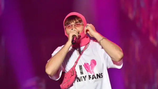 Mata raper w różowej kominiarce i okularach, z torbą na ramię i koszulką "I MY FANS", śpiewa na koncercie.
